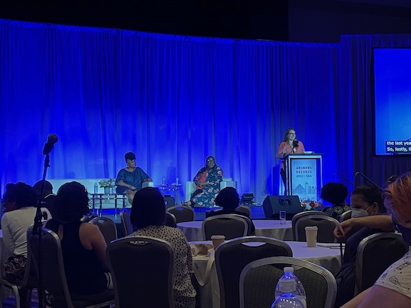 Photograph of the keynote address stage at the Sheraton Boston with three women, two sitting and one standing at the podium.